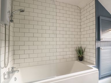 A bathroom with a bathtub shower combo white subway tile walls a small potted plant and blue louvered cabinets at Haven in Porthleven