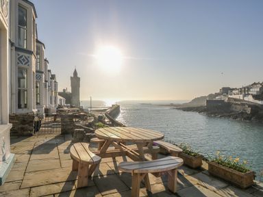 An outdoor seating area with wooden tables and benches overlooking a harbor with a clock tower and buildings at Haven in Porthleven