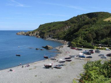 A pebble beach with boats on shore cars parked nearby and hills covered in vegetation at Cockle Island Cottage in Porthallow