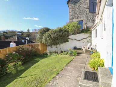 A garden with a lawn a stone path and two chairs by a white building at Cockle Island Cottage in Porthallow