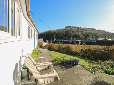 A backyard with two plastic chairs potted plants a lawn and a wooden fence with a hill in the background at Cockle Island Cottage in Porthallow