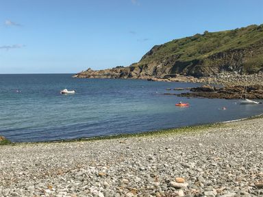 A rocky beach with a small boat and a kayak on the water with a hill in the background at Cockle Island Cottage in Porthallow