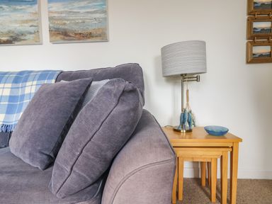 A sofa with cushions and a blue check blanket next to a wooden nesting table with a lamp and bowl at Cockle Island Cottage in Porthallow