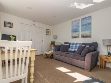 A living room with a grey sofa a table with white chairs and paintings on the wall at Cockle Island Cottage in Porthallow