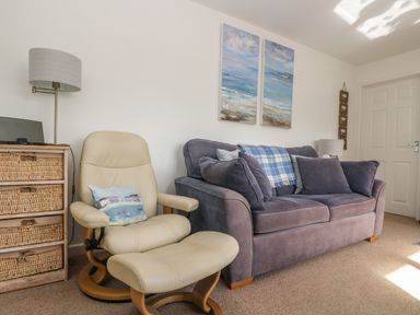 A living room with a gray sofa a beige armchair with a matching footrest a lamp and wicker drawers at Cockle Island Cottage in Porthallow