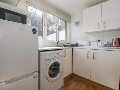 A kitchen with white cabinets a washing machine a refrigerator a microwave toaster and a kettle at Cockle Island Cottage in Porthallow