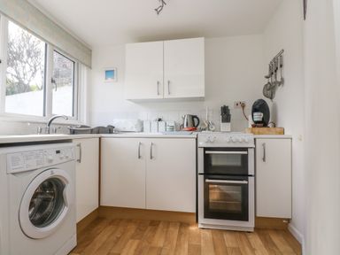 A kitchen with white cabinets a washing machine an oven a kettle and a coffee machine at Cockle Island Cottage in Porthallow