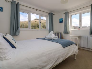 A bedroom with a bed folded towels on it two windows with curtains a side table and a chair at Cockle Island Cottage in Porthallow