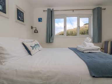 A bedroom with a bed with white sheets and blue blanket with folded towels near a window with blue curtains at Cockle Island Cottage in Porthallow
