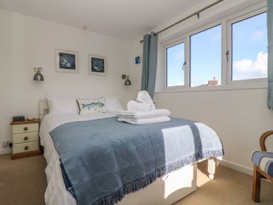 A bedroom with a bed covered in white and blue linens next to a small wooden bedside table and a window with blue curtains at Cockle Island Cottage in Porthallow