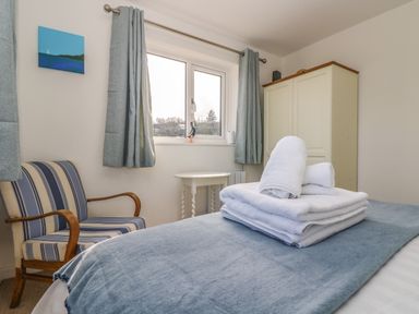 A bedroom with a bed covered by a blue blanket and white towels on top a striped armchair a window with blue curtains a small white side table and a cream wardrobe at Cockle Island Cottage in Porthallow