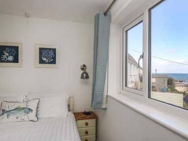 A bedroom corner with a bed, bedside table, wall lamp, framed pictures, and a window showing a sea view at Cockle Island Cottage in Porthallow