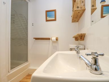 A bathroom with a sink two taps a shower cubicle wooden shelves and a toilet paper holder at Cockle Island Cottage in Porthallow