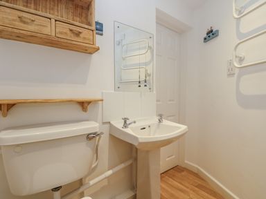 A small bathroom with a toilet and pedestal sink under a wooden shelf and cabinet at Cockle Island Cottage in Porthallow