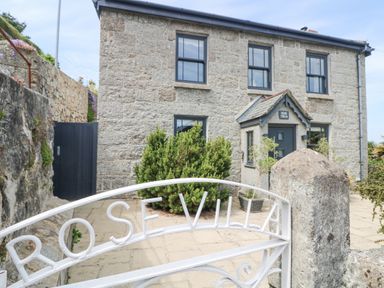 Stone house with black framed windows and a small porch behind a white metal gate with rose villa on the gate at Rose Villa in Newlyn