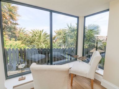 A corner seating area with two upholstered chairs by large windows overlooking palm trees at Rose Villa in Newlyn