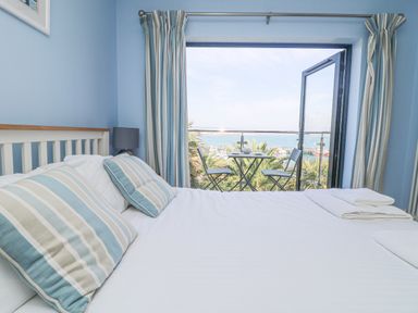 A bedroom with bed and striped pillows next to an open door leading to a balcony with chairs and table overlooking the sea at Rose Villa in Newlyn
