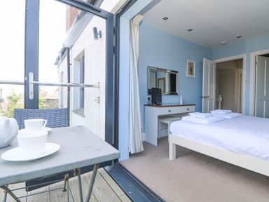 A bedroom with a bed and towels on it and a desk with a mirror and a small tv next to a balcony with a table and cups at Rose Villa in Newlyn
