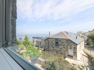 A view of stone buildings trees and the sea seen through a window at Rose Villa in Newlyn