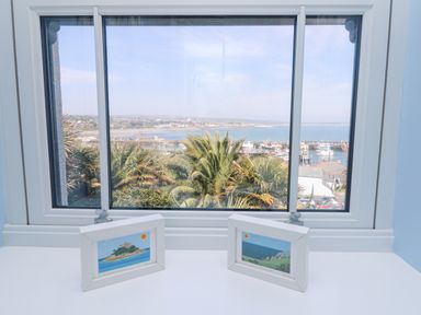 A window with a view of palm trees boats and water with two framed pictures on the windowsill at Rose Villa in Newlyn