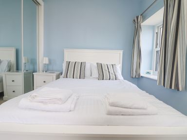 A bedroom with a white bed with striped pillows two bedside tables with lamps and a window with striped curtains at Rose Villa in Newlyn