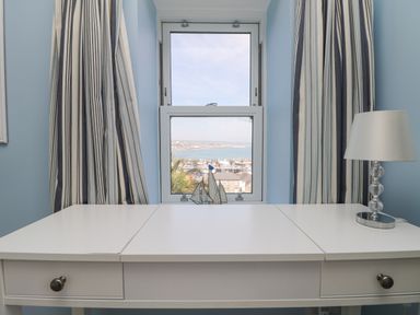 A white desk with drawers a lamp and striped curtains by a window overlooking a harbor at Rose Villa in Newlyn