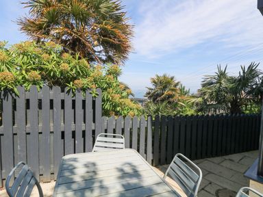 An outdoor patio with a metal table and chairs facing a grey wooden fence with trees and a view of the sea at Rose Villa in Newlyn