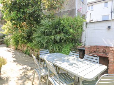 An outdoor patio with a metal table and six chairs near a brick barbecue and green plants at Rose Villa in Newlyn