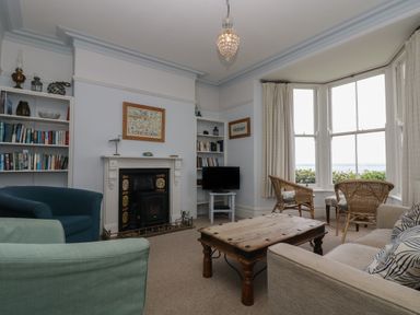 A living room with a fireplace bookshelves armchairs a wooden coffee table a sofa and a bay window with wicker chairs at Lookout in Mousehole
