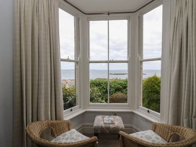 A seating area with two wicker chairs and a patterned ottoman by a bay window overlooking greenery and the sea at Lookout in Mousehole