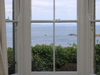 A window view of the sea with a small boat and greenery outside at Lookout in Mousehole