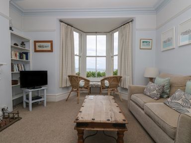 A living room with a sofa and cushions a wooden coffee table two wicker chairs by a bay window a TV on a small table and bookshelves at Lookout in Mousehole