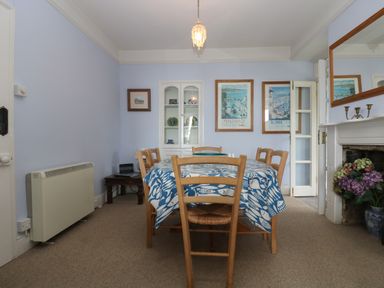 A dining room with a wooden table and chairs a blue and white tablecloth framed pictures a fireplace with flowers and a wall cabinet at Lookout in Mousehole