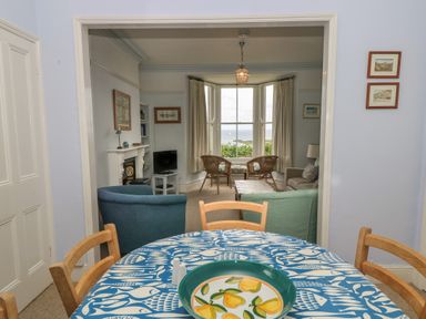 A dining area with a round table and chairs facing a living room with armchairs a sofa a coffee table and a bay window at Lookout in Mousehole