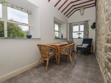 A dining area with a wooden table and wicker chairs next to windows with a stone wall and a blue armchair at Lookout in Mousehole