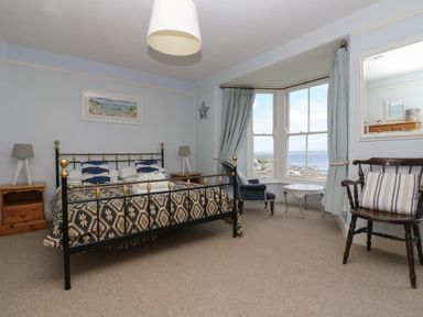 A bedroom with a metal bed frame two bedside tables with lamps a blue armchair a side table a wooden chair and a bay window showing a coastal view at Lookout in Mousehole