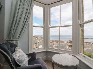 A corner with a blue armchair with a patterned cushion next to a round wooden table by large windows overlooking rooftops and the sea at Lookout in Mousehole