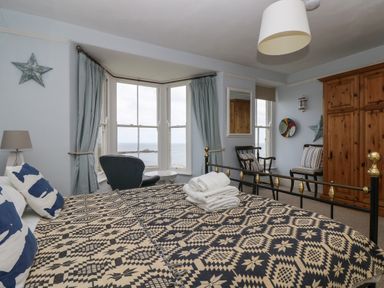 A bedroom with a patterned bedspread a bay window with a view of the sea chairs and a wooden wardrobe at Lookout in Mousehole