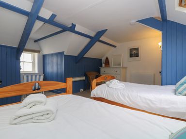 A bedroom with two single beds, blue wooden beams and paneling, a chest of drawers, and a wicker chair at Lookout in Mousehole