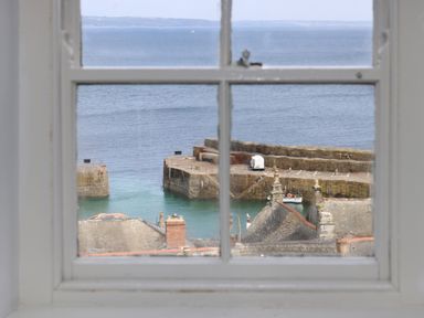 A sea view through a window showing a stone pier a white van rooftops and distant land at Lookout in Mousehole