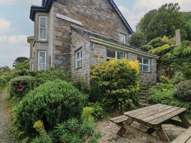 A stone house surrounded by bushes and greenery with a wooden picnic table in front at Coronation Villas in Mousehole