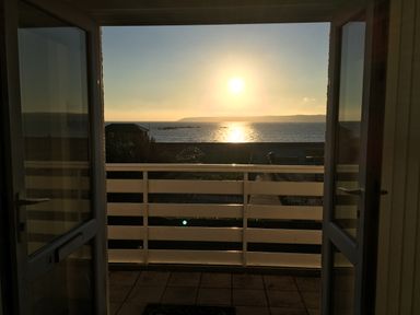 A view of the sun setting over the sea from a balcony with white railings and open glass doors at Sea Breeze in Marazion