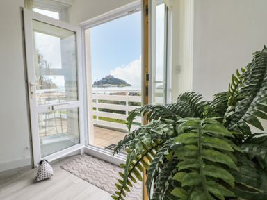 An open glass door leading to a balcony with a view of a hill and sea with a large green plant inside at Sea Breeze in Marazion
