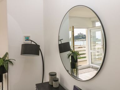 A round wall mirror reflecting an open door with a view of a castle across water beside a floor lamp plant and small containers at Sea Breeze in Marazion