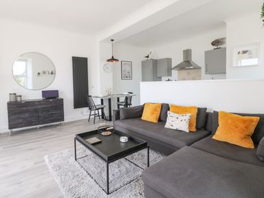 A living room with a gray sectional sofa and yellow pillows a black coffee table with decor a dining table with two chairs and a kitchen with gray cabinets at Sea Breeze in Marazion