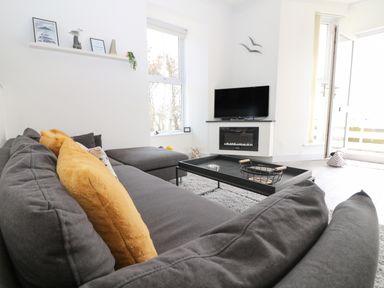 A living room with a gray sectional sofa with yellow pillows a black coffee table a flat screen tv mounted above a fireplace and windows with a door at Sea Breeze in Marazion