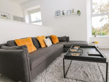 A living room with a gray sectional sofa with orange and patterned cushions a black tray table with decorative items a gray rug and a large window at Sea Breeze in Marazion