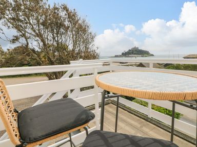 A balcony with a glass table and two chairs overlooking a tree and an island with a castle at Sea Breeze in Marazion
