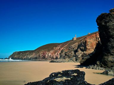 A sandy beach with rocky cliffs and old stone buildings on the hill at Mithian Cottage in St Agnes