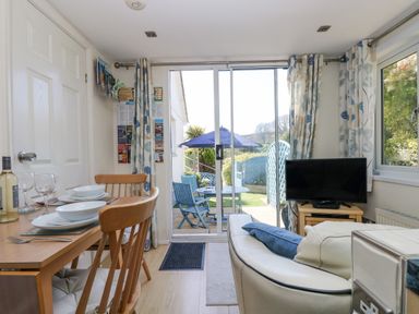 A dining area with wooden chairs and table set with dishes next to a living area with white sofa and television by sliding glass doors to a patio with blue chairs and table at Respryn Mevagissey